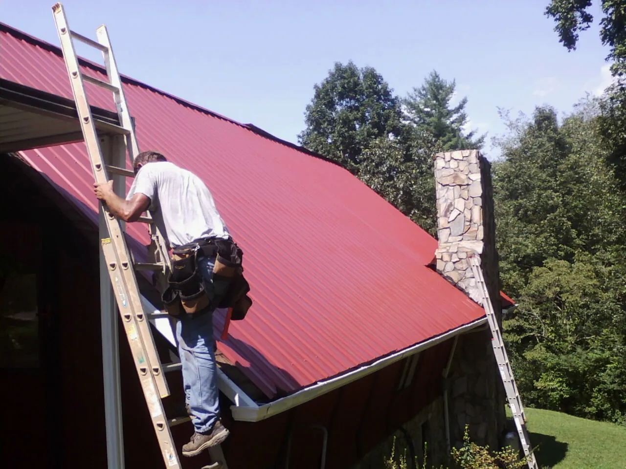 Roofer on a ladder beside a red metal roof and stone chimney — Soffit & Fascia Repair by Roofing Brothers in Magazine